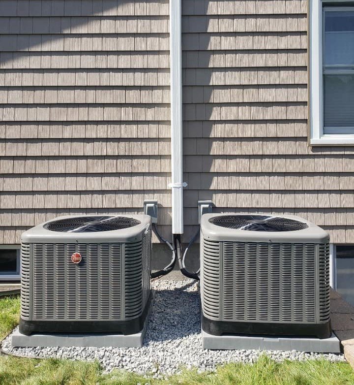 Dual Rheem HVAC condensers installed outside a house with vinyl shake siding, placed on concrete pads over gravel for stability and efficient cooling performance.